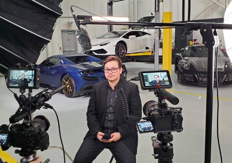 Juan on the set of Supercar Saints, seated for an interview in front of cameras, with supercars and his Porsche behind him